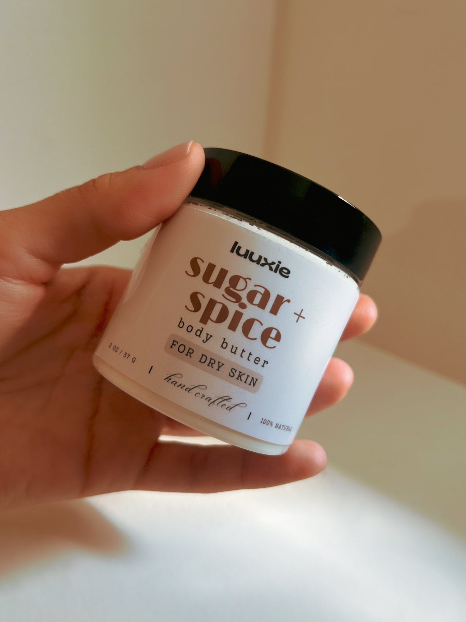 Hand holding a jar of Luuxie sugar + spice body butter against a neutral background